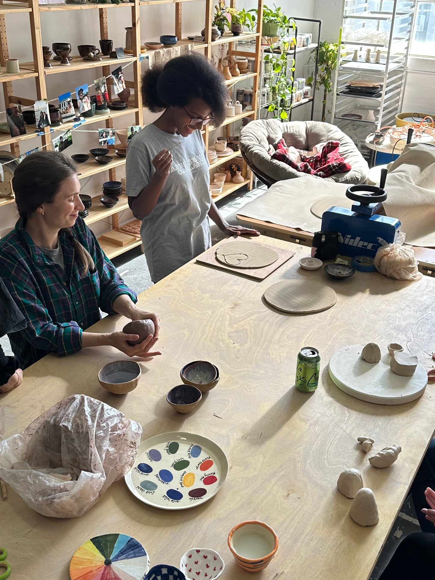 Two people working on pottery in a studio with shelves and a couch in the background.